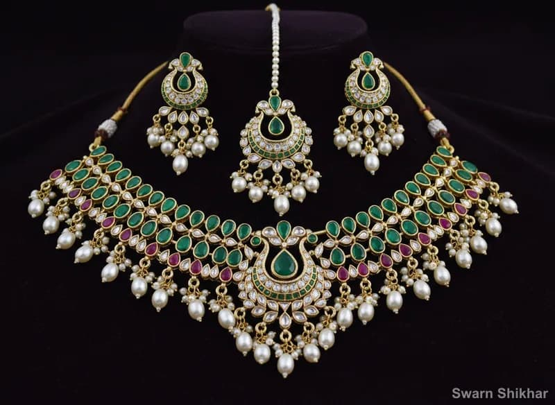 Bridal necklace set in 22K gold with matching earrings, red and green stones and pearl drops on maroon velvet at Swarn Shikhar Jewellers Chembur